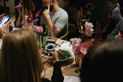 Menschen sitzen bei einem Dinner zusammen, eine Frau schießt ein Foto von ihrem Essen